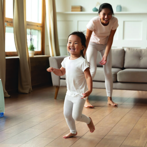 Full length overjoyed little preschool cute vietnamese ethnic baby girl running barefoot on warm floor, having fun playing with energetic young asian mother or nanny, leisure daycare activity.